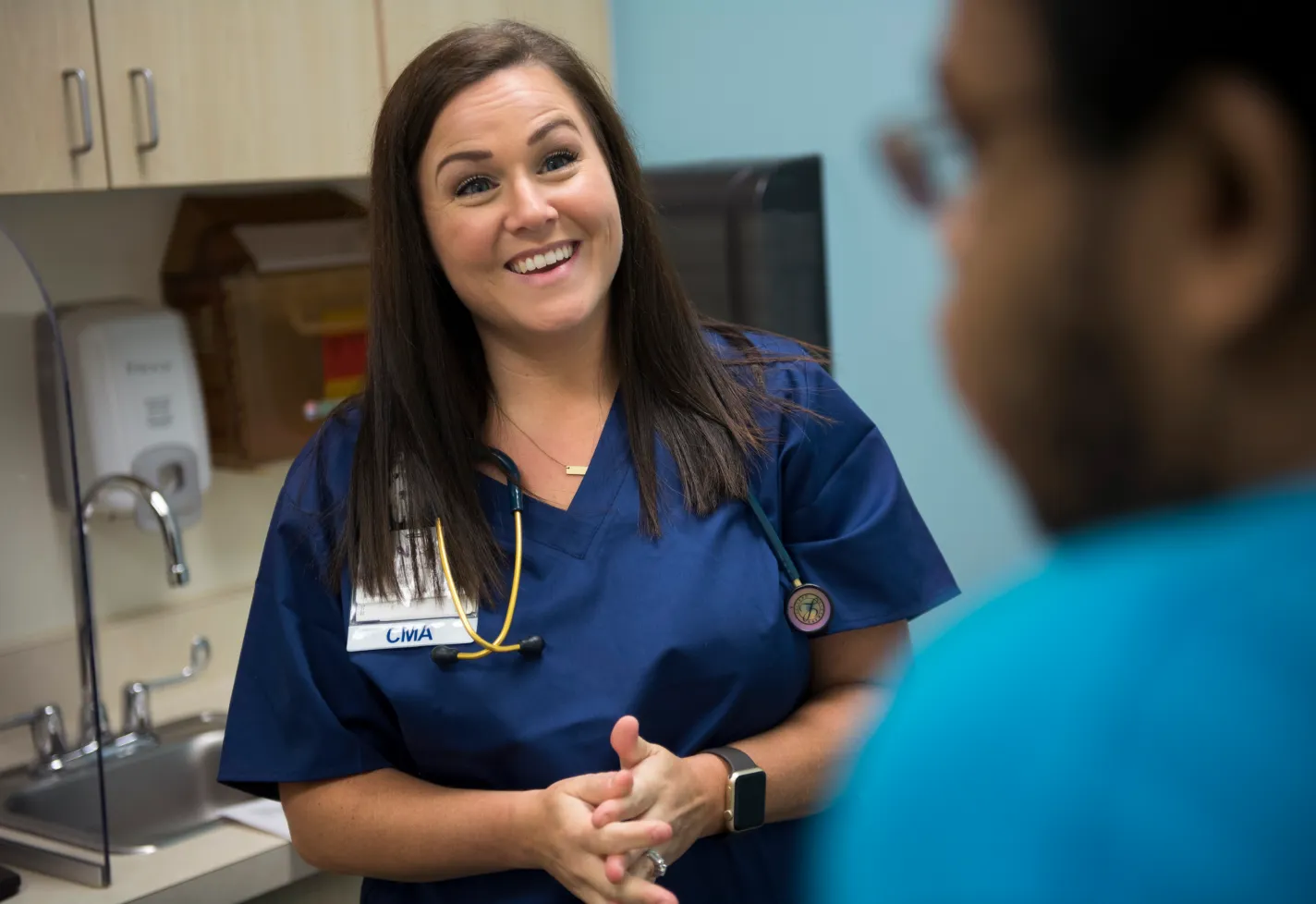 A Novant Health Certified Medical Assistant (CMA) is in an exam room talking and smiling with a male patient. 