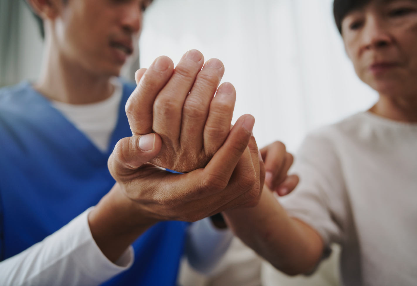 A caregiver holds the hand of a senior woman, checking her wrist. The room is bright.