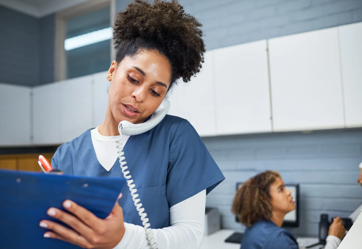 A female healthcare worker takes notes while talking on a phone.