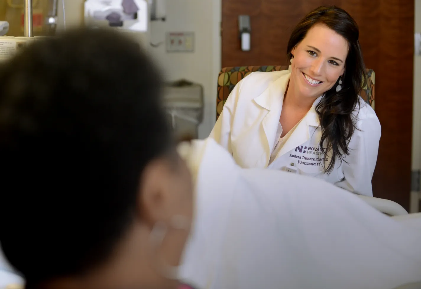 Novant Health pharmacist, Dr. Demera, is sitting at the bedside of a patient talking and smiling. 