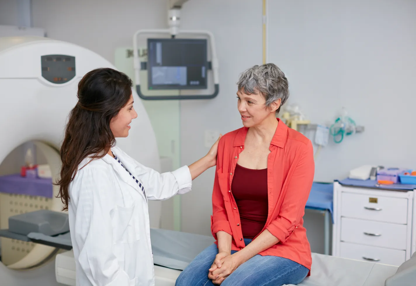 A healthcare provider is resting her hand on a patient's shoulder as she is talking and preparing her for an imaging scan. 