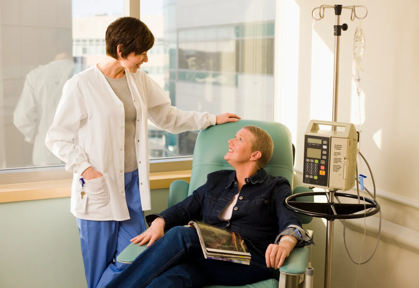 A woman is sitting in a chair receiving infusion treatment while talking with her healthcare provider. 
