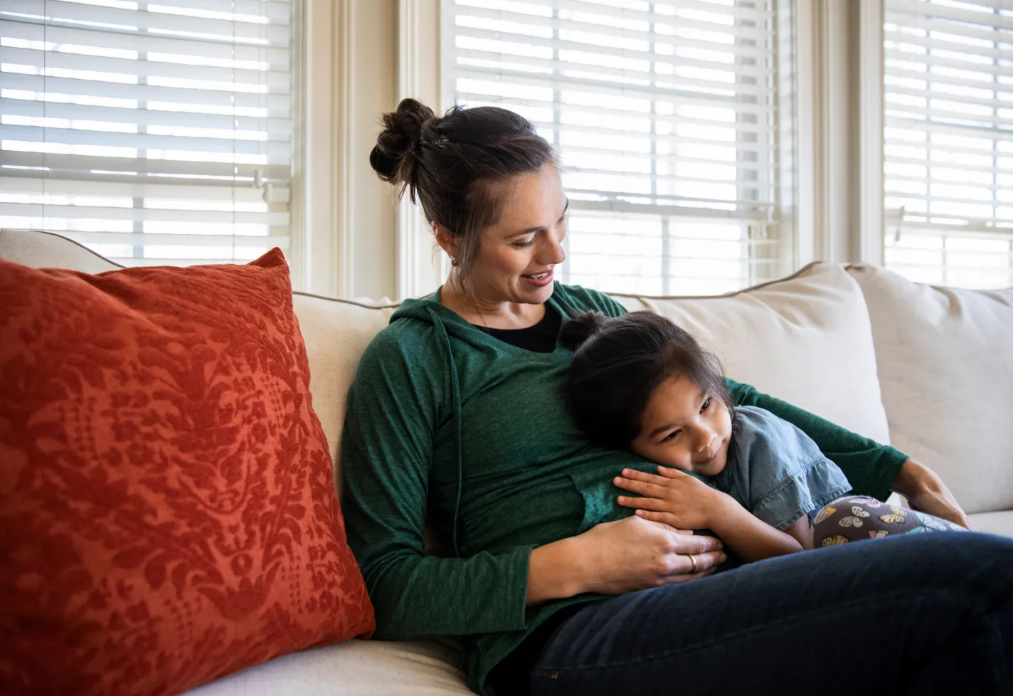 A mother and her young child are hugging each other as they sit on the couch. The child has their hand and ear on their mothers abdomen.