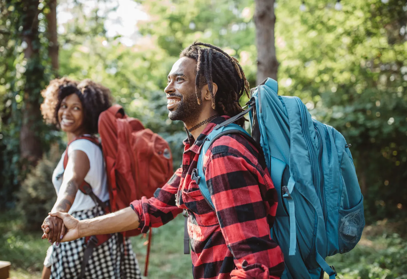 An African American couple is out wearing camping backpacks and holding hands as they hike together. 