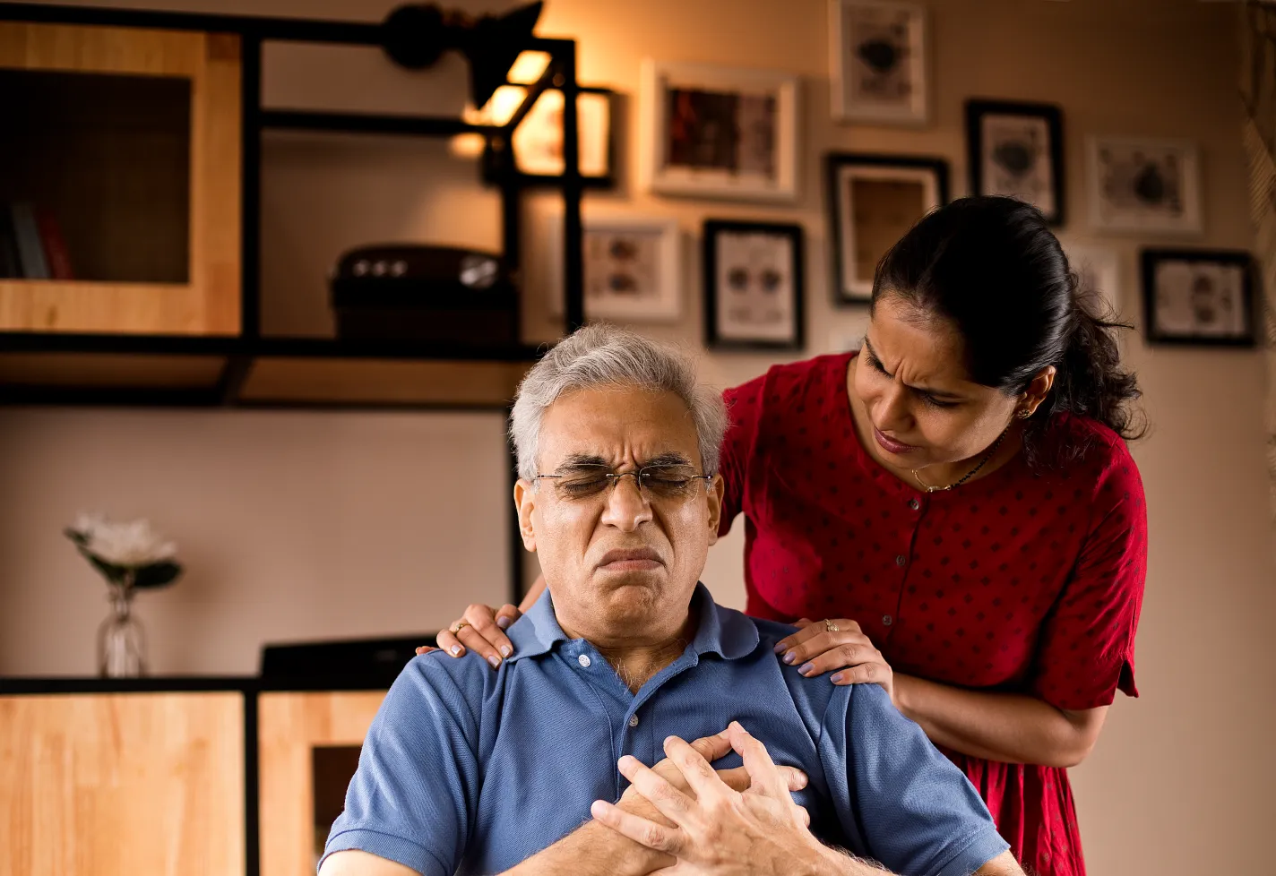 A senior man is sitting and holding his chest. His wife is standing behind him concerned. 