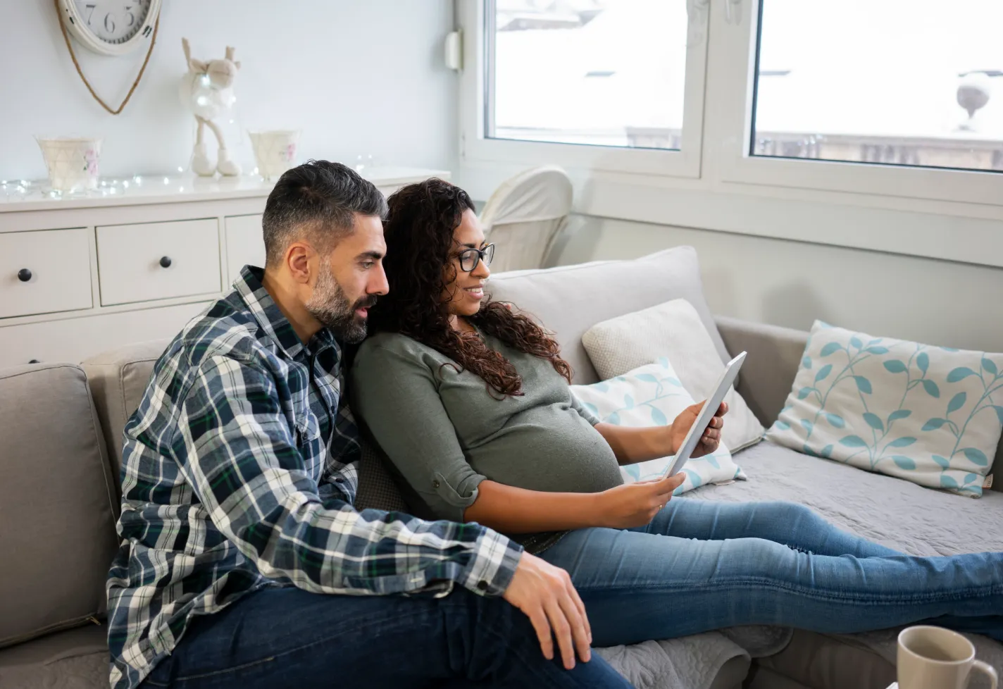 A man and his pregnant partner are sitting on their couch together reviewing information on a tablet. 