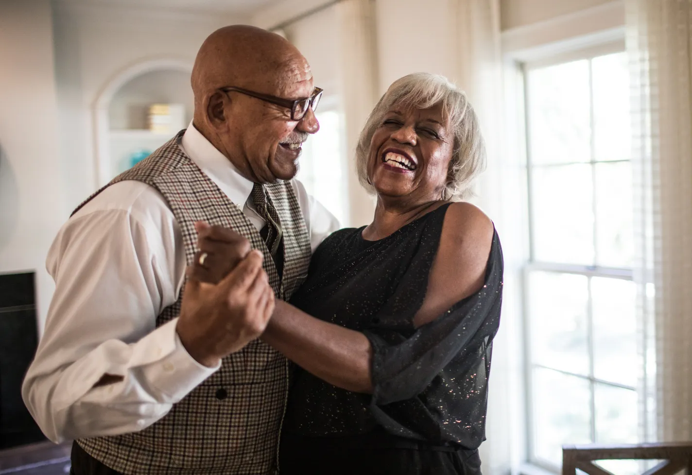 A senior, African American, couple are holding hands, dancing, and smiling. 