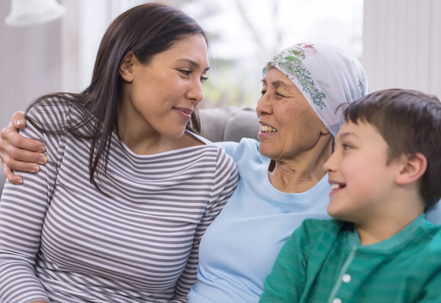 A grandmother is wearing a headscarf as she hugs and smiles at her adult daughter and grandchild. 