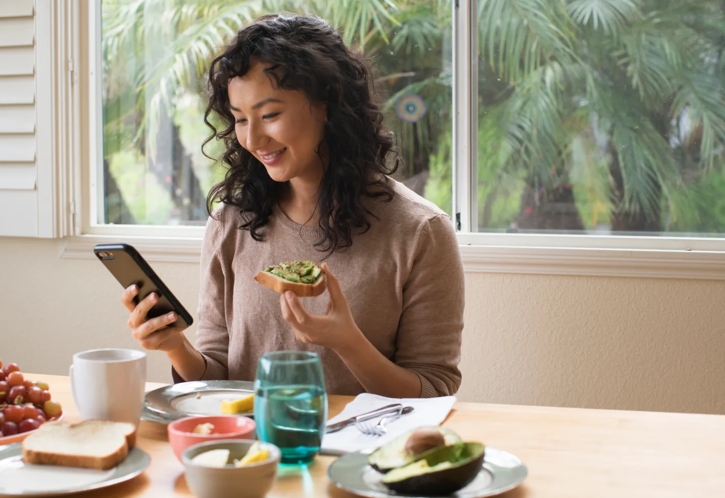 A young woman is sitting at the kitchen table eating avocado toast while looking at her smartphone. 