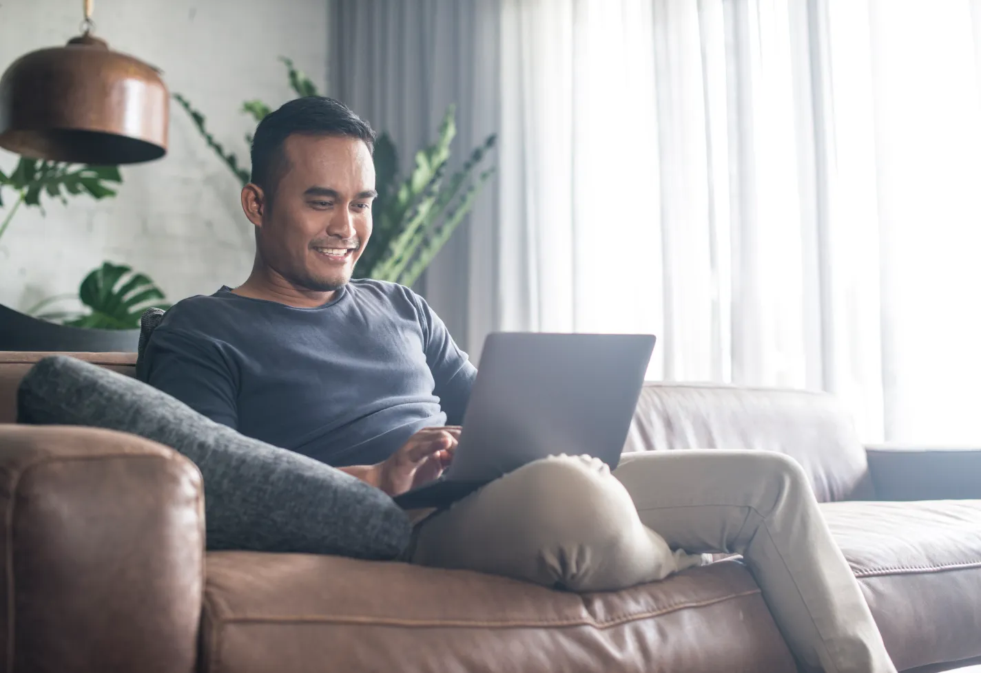 A man smiles while sitting on a couch and using a laptop.