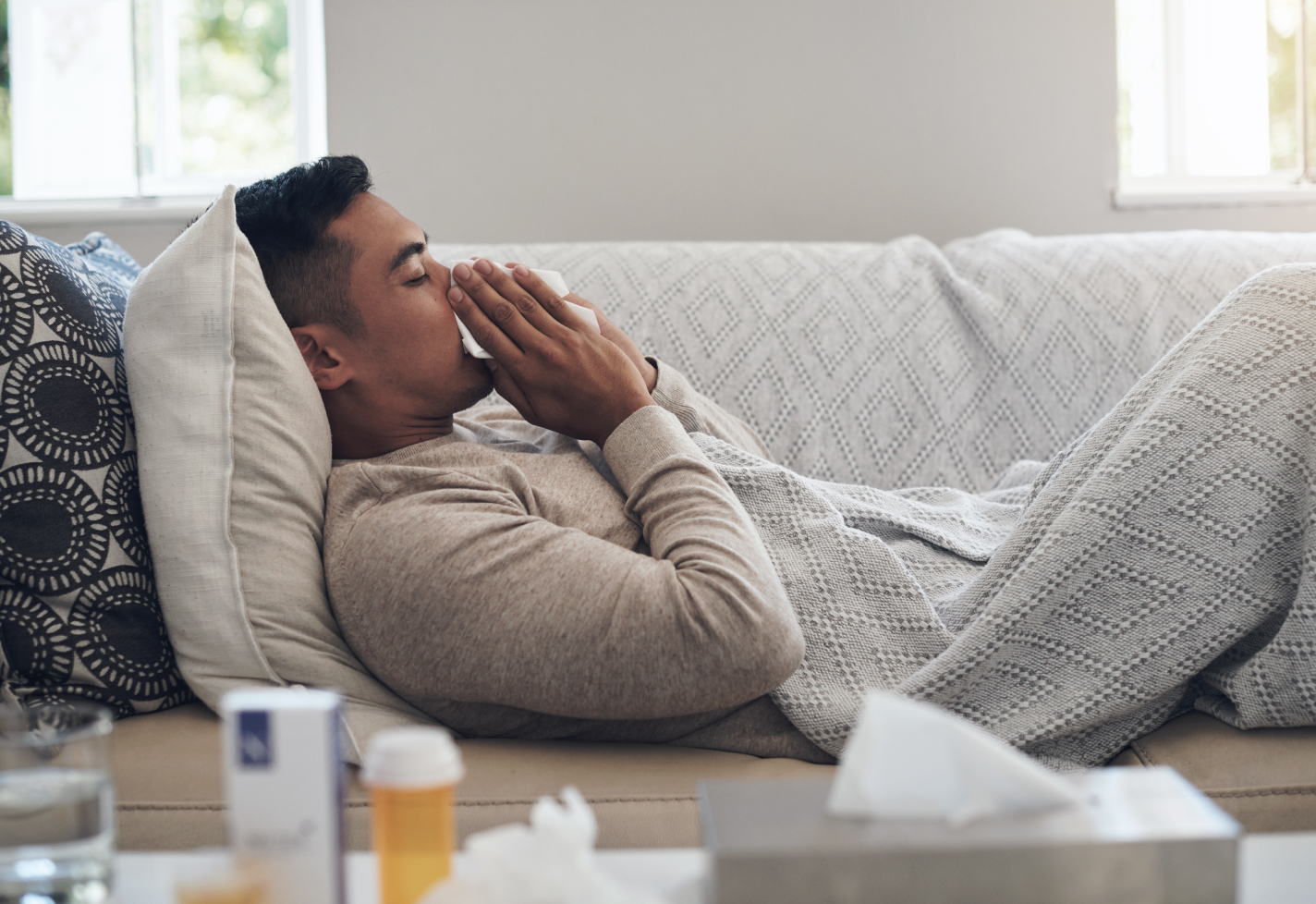 A man laying on a couch while blowing his nose with a tissue.