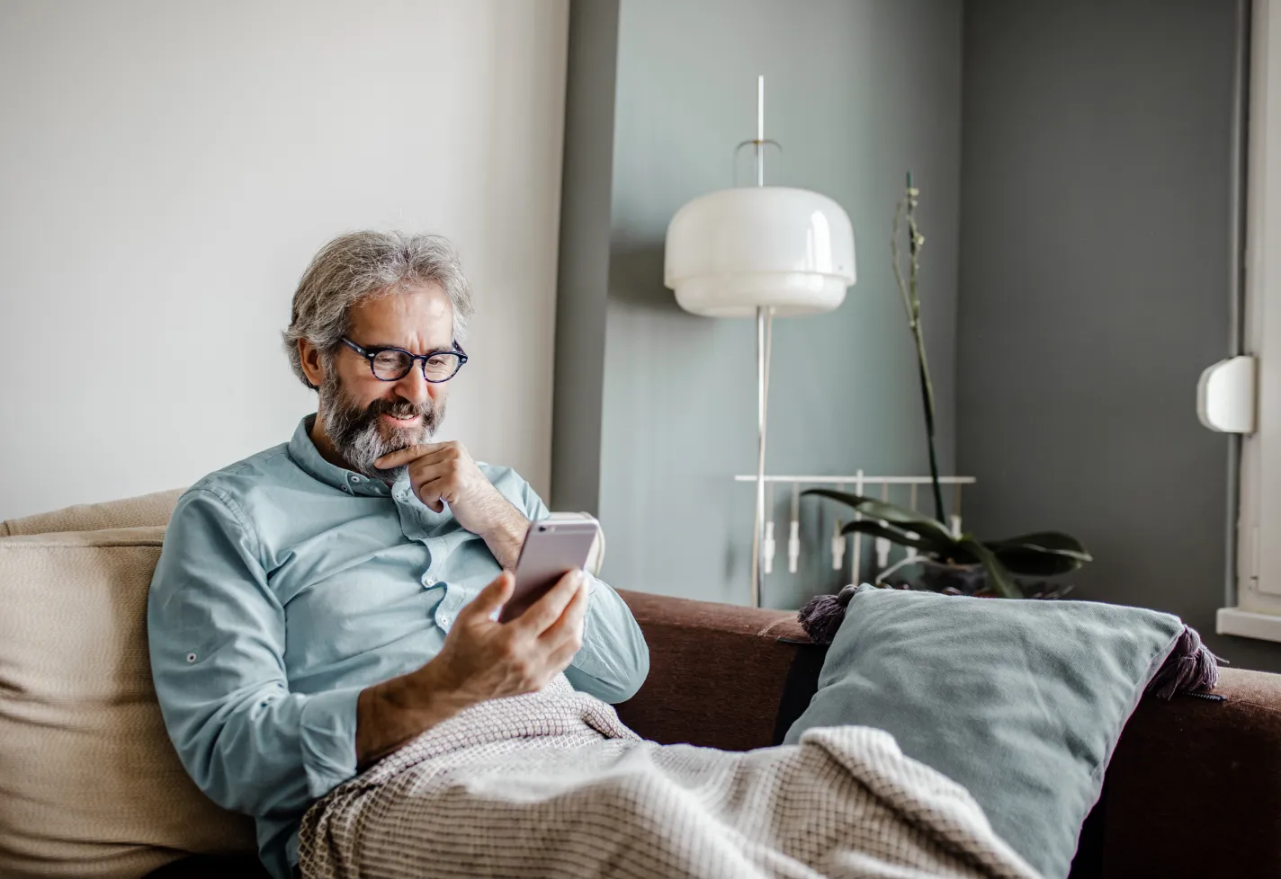 A man is sitting on the couch in his home as he smiles and reads information on his smart phone. 