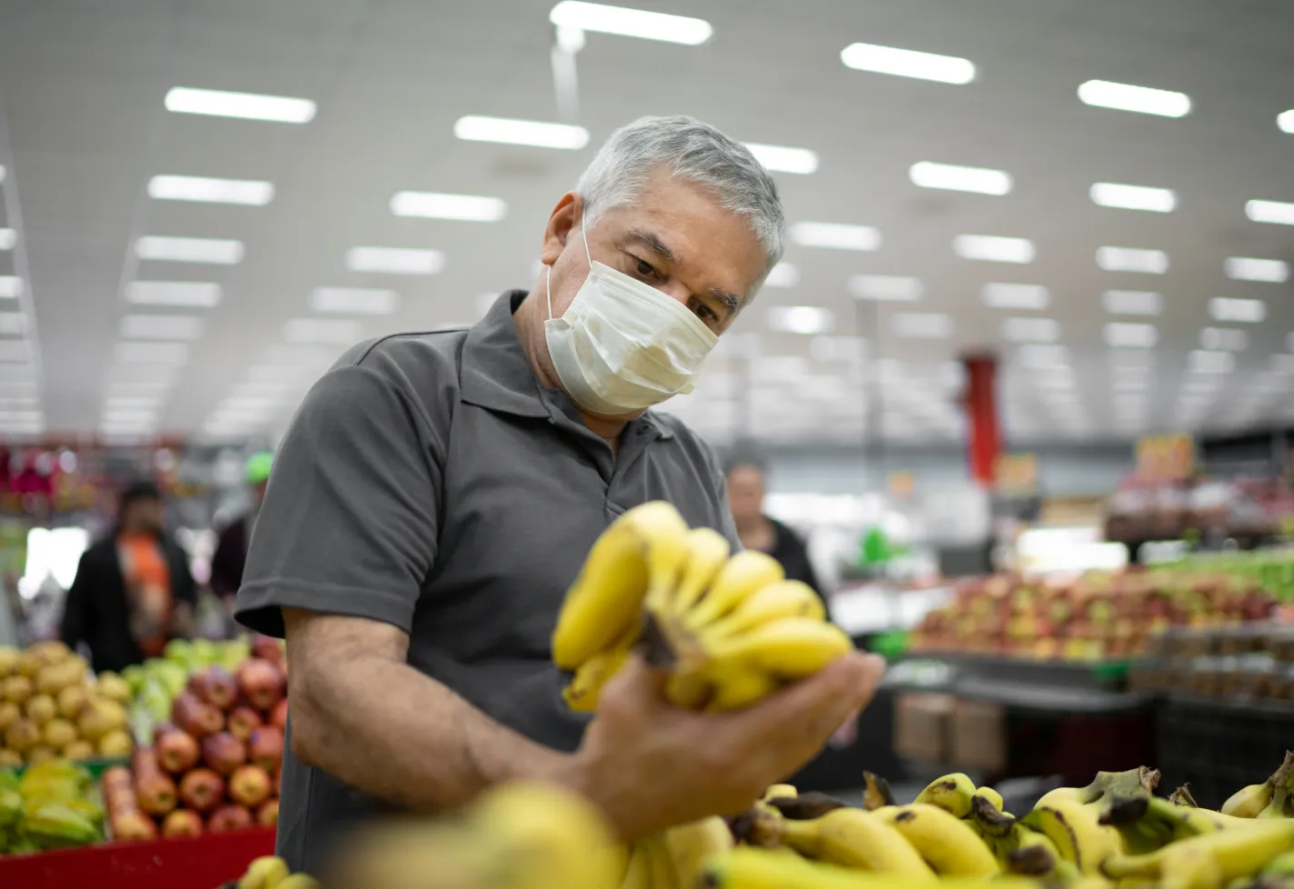 Senior man with disposable medical mask shopping in supermarket holding bananas.