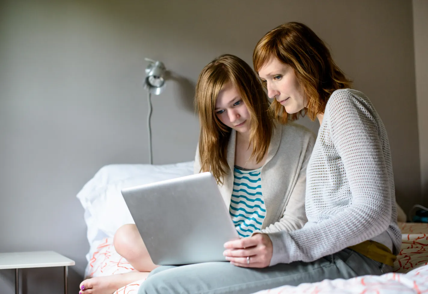 A mother and daughter are sitting on a bed reading information on a laptop. 