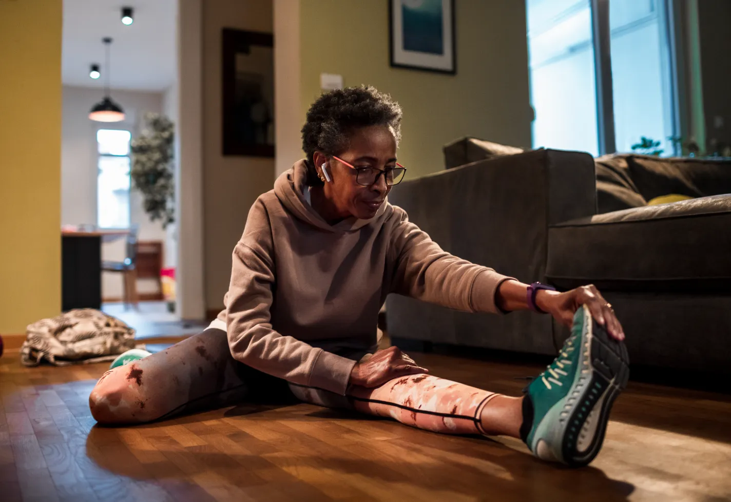 A senior woman is sitting on the floor, stretching, as she prepares for a run. 