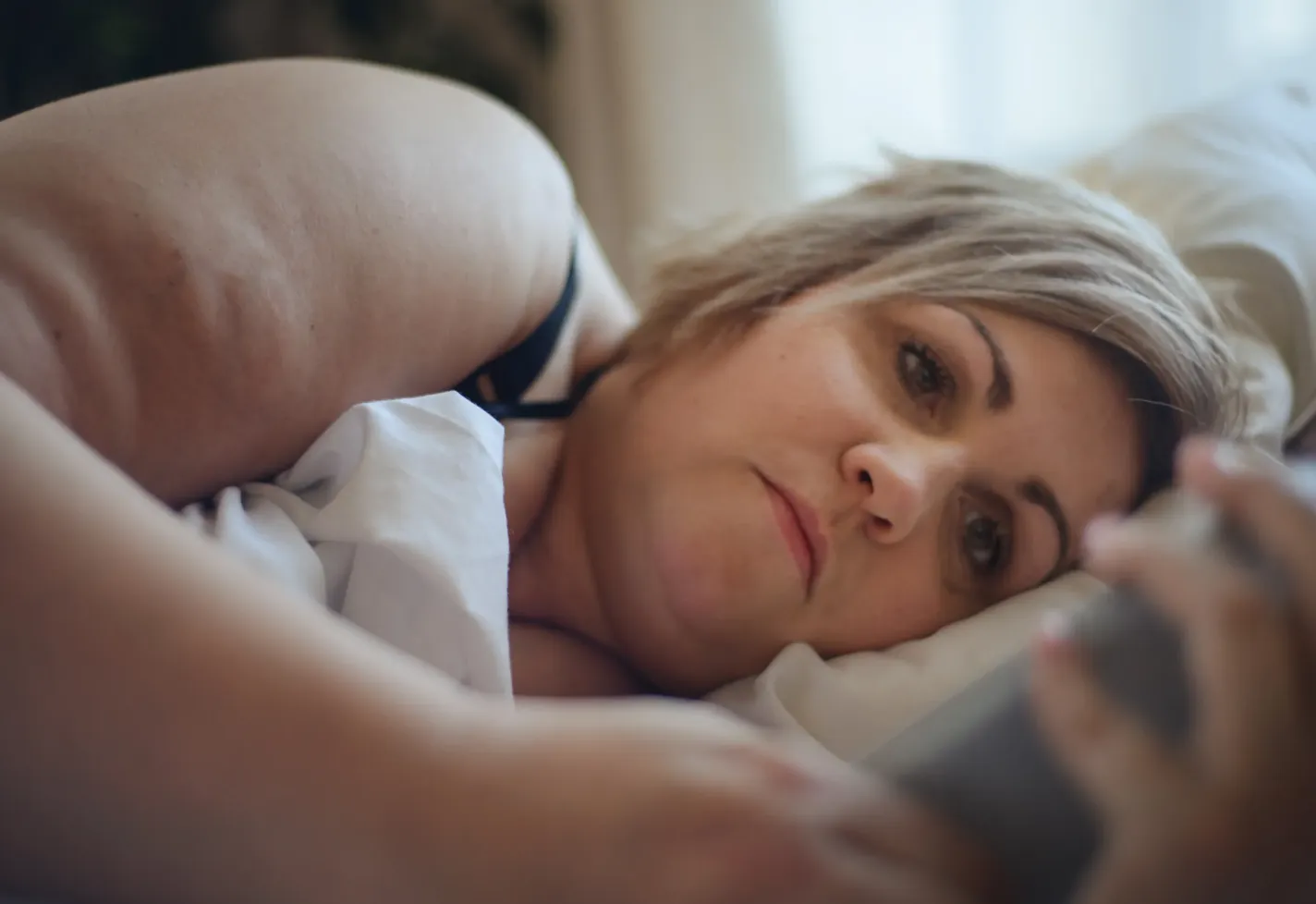 A woman is laying in bed, looking at information on her smartphone. 