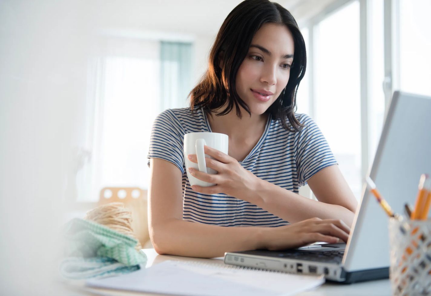 A woman holding a coffee mug while typing on a laptop and sitting at a desk.