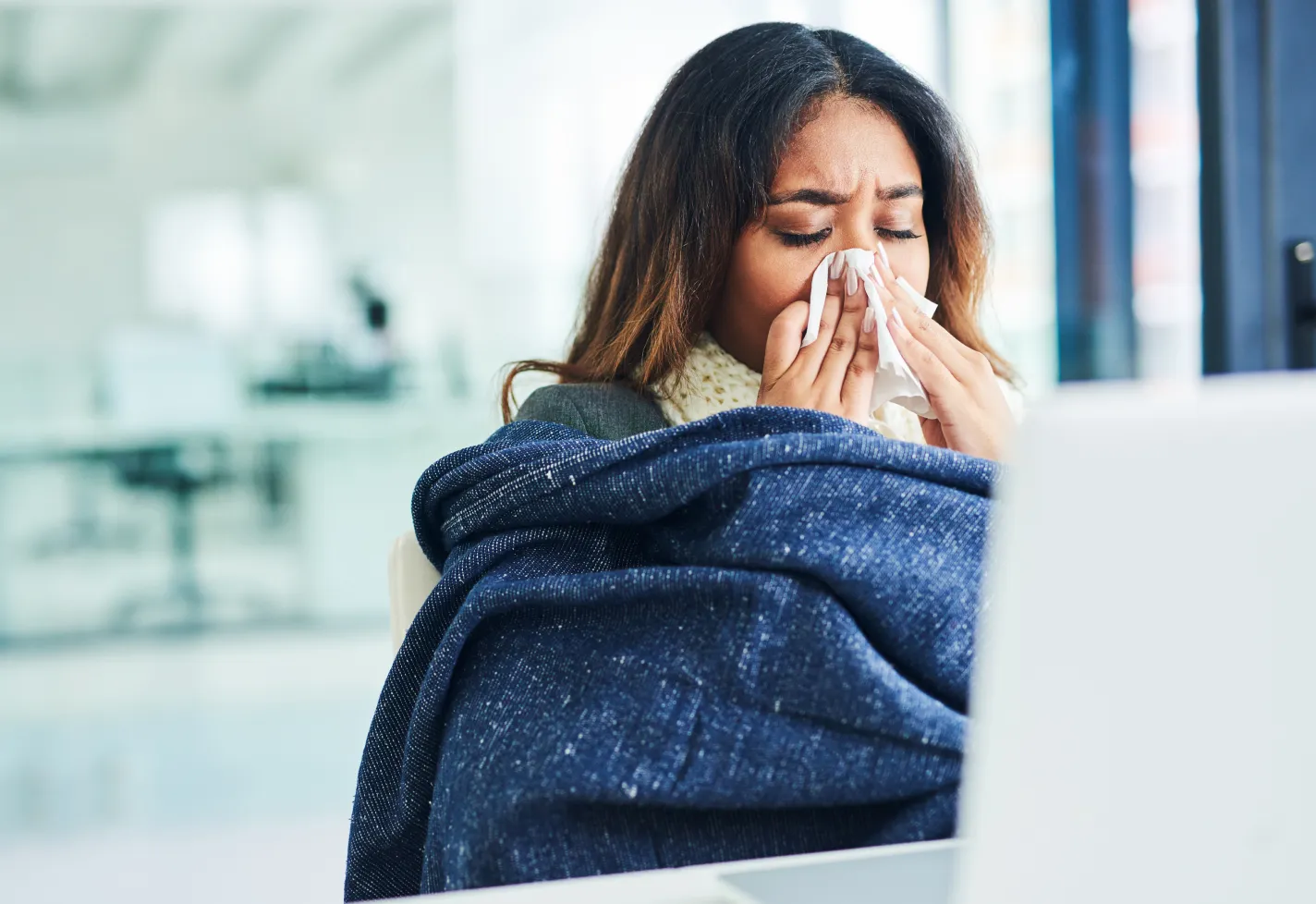A woman covers her nose with tissue while using a laptop.