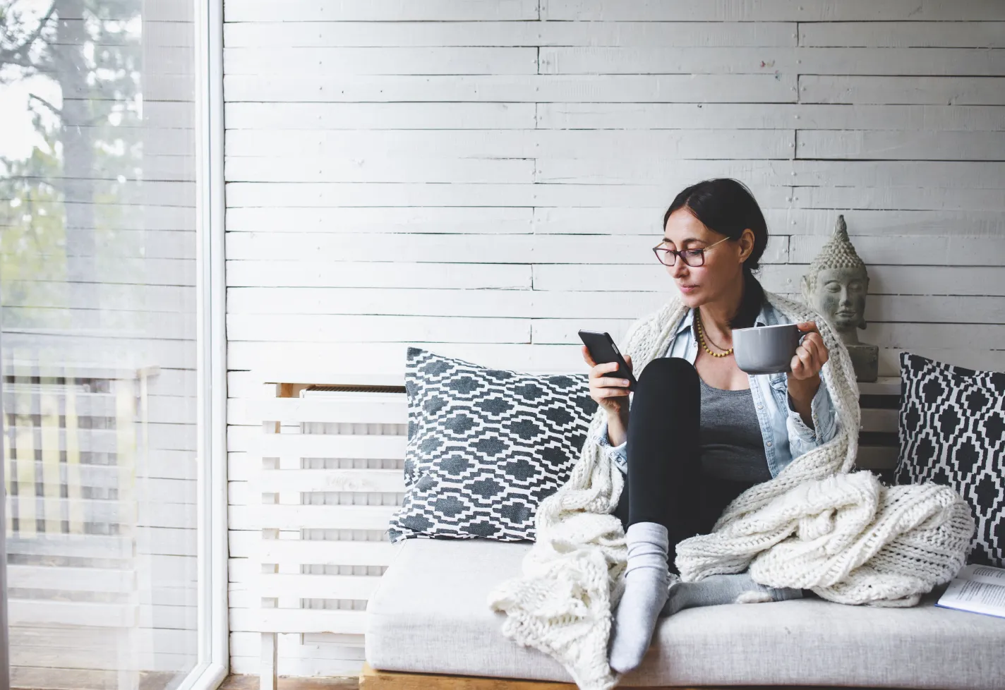 A woman is sitting on the couch, wearing a blanket, drinking coffee while on her smartphone. 