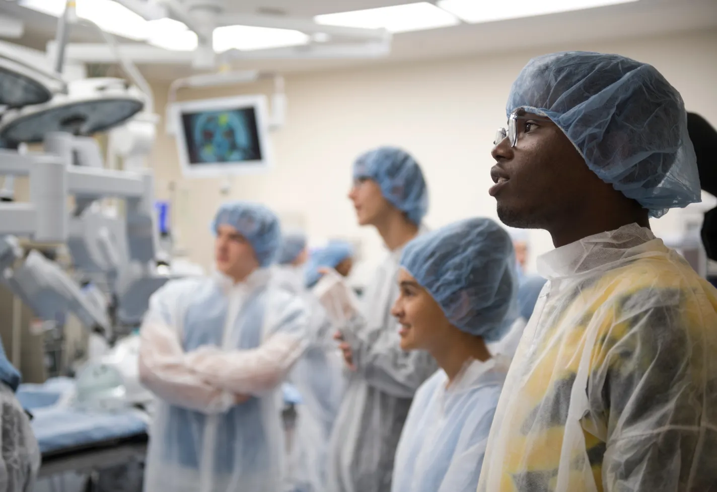 A group of students are wearing personal protective equipment (PPE) as they stand and observe in an operating room.  