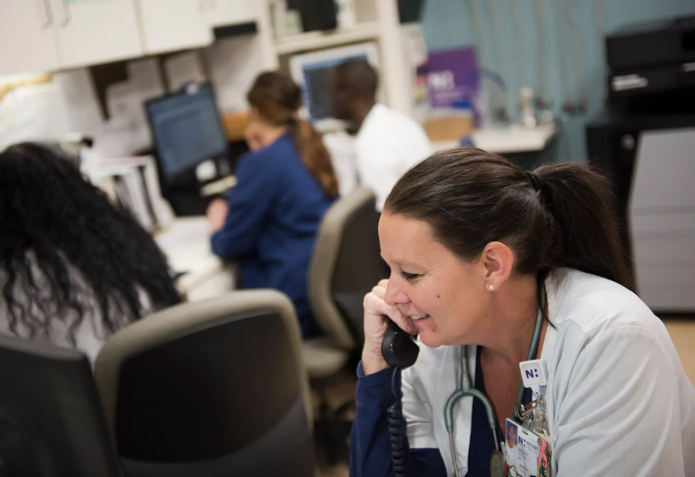 A Novant Health nurse is sitting at a nurses station, in front of a computer, talking on the phone. 