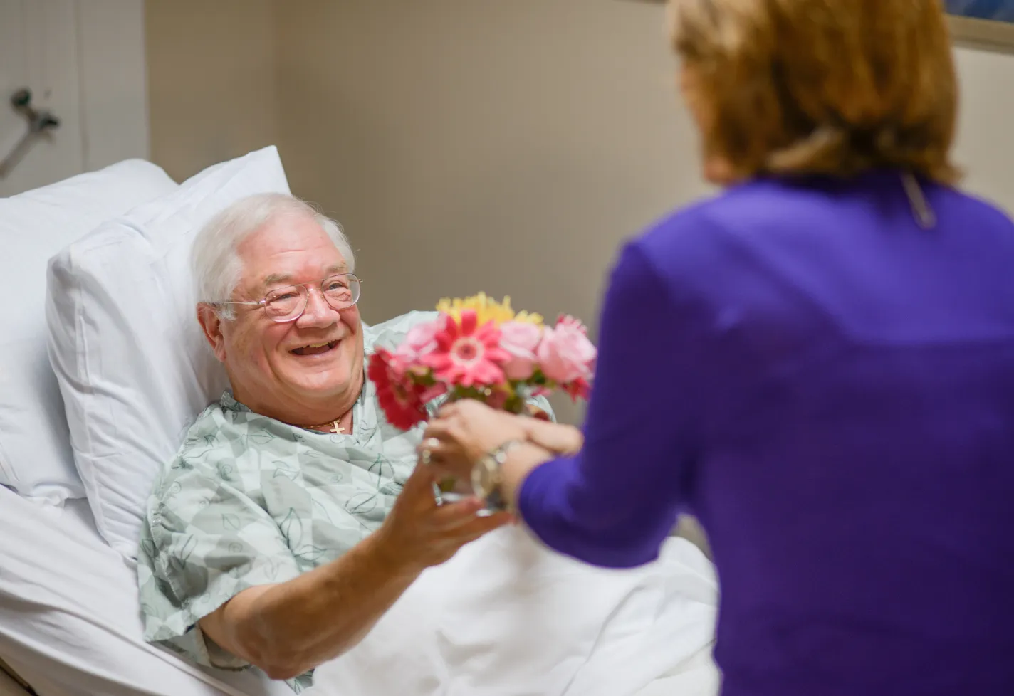 A Novant Health volunteer is delivering flowers to a patient who is lying down in a hospital bed.