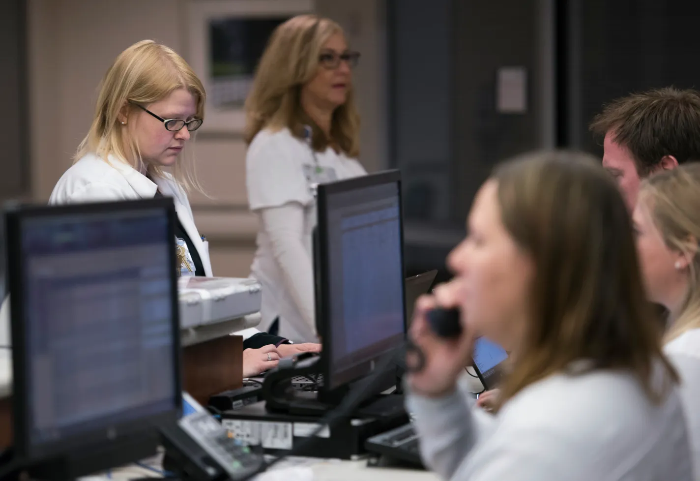 Several team members are surrounding a nurses station. Most team members are typing computers, while one is on the phone. 