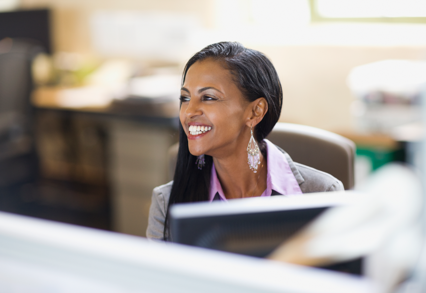 A woman in a gray blazer and pink blouse smiling while seated at a desk with computers.