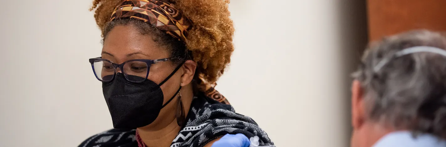 A woman, who is wearing a mask, is in an exam room receiving a vaccination from her Novant Health doctor. 