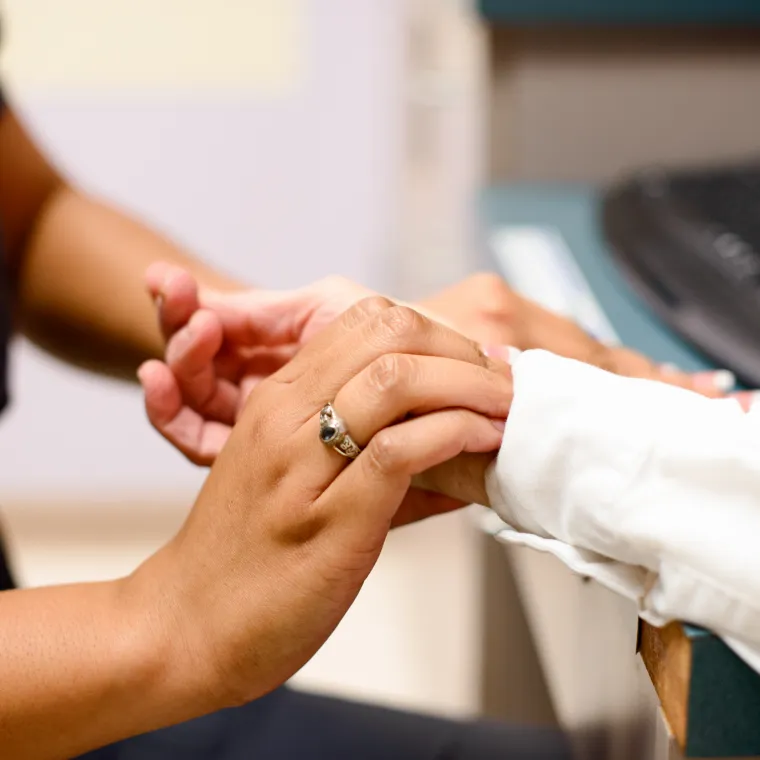 Nurse and patient are sitting across from each other in a clinic exam room. The nurse holds and positions the patients hands to check for their pulse. 