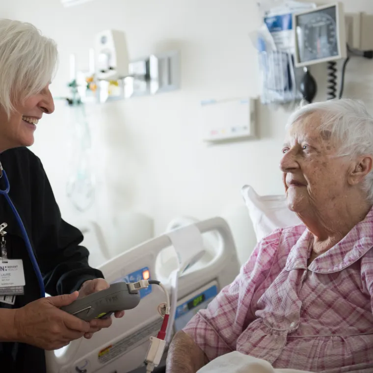 A Novant Health nurse is at the bedside of a senior patient. The nurse is smiling and talking with the patient as she holders a large medical devce in her hand. 