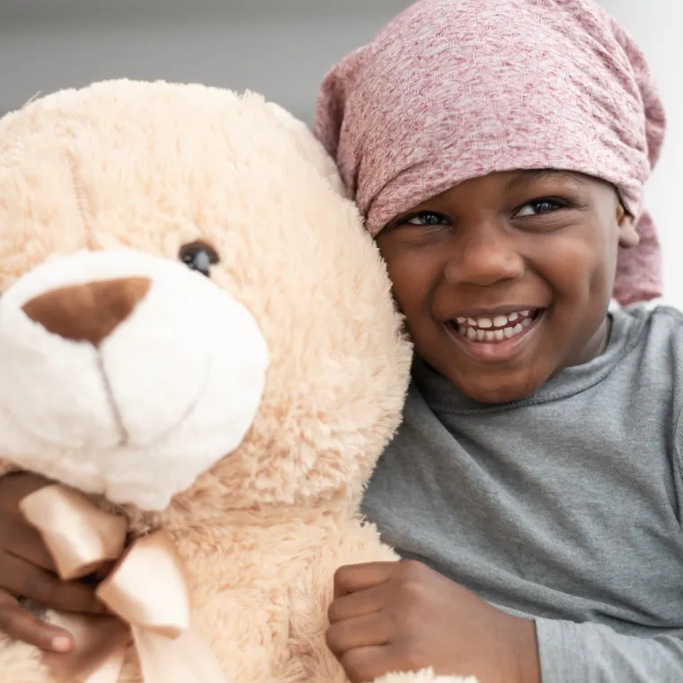 Young child wearing is wearing a head wrap and hugging a teddy bear. 