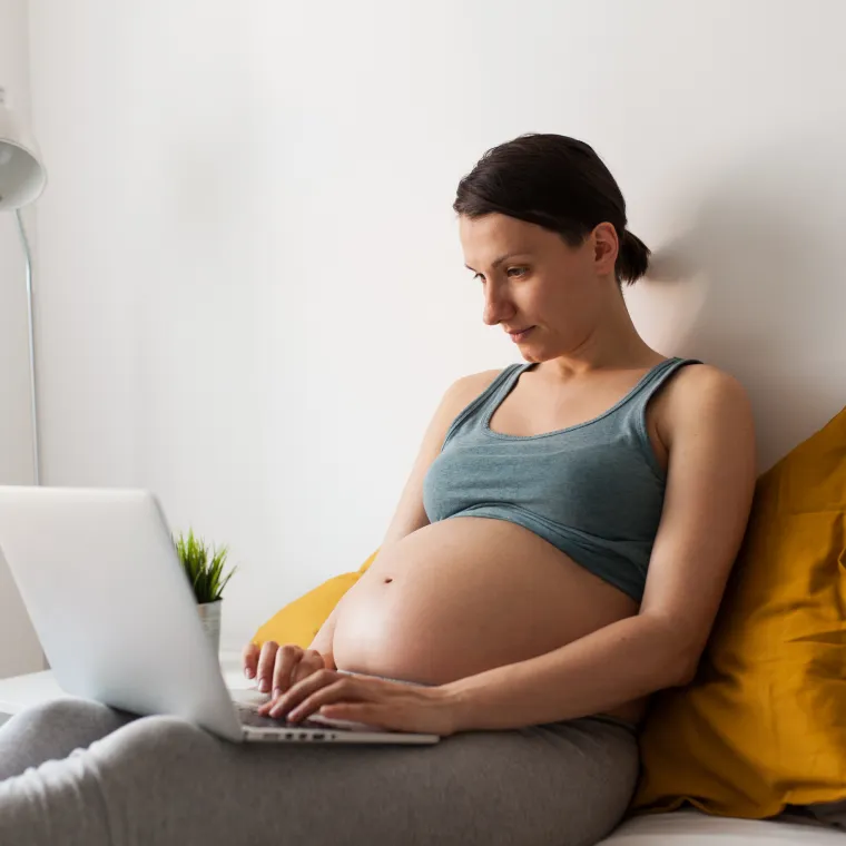 A pregnant woman typing on a laptop in her bedroom.
