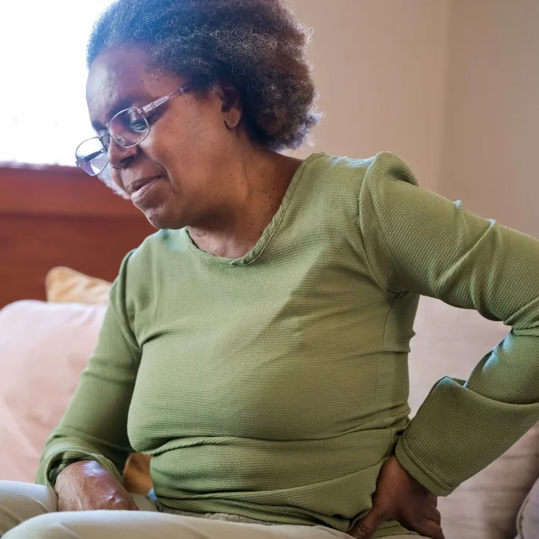 Senior woman sitting on couch holding her back due to pain. 