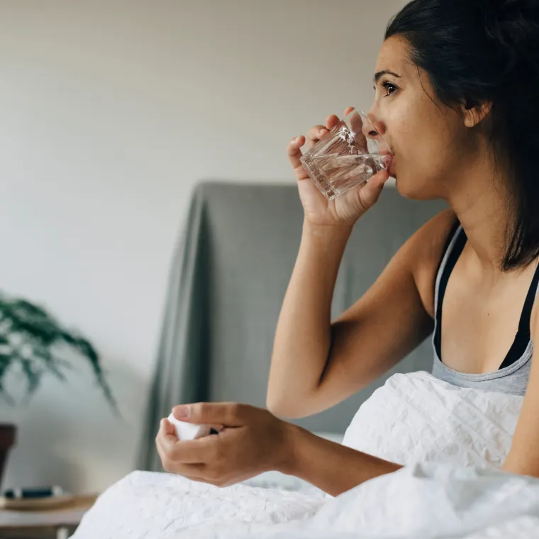 Woman sitting in bed drinking a glass of water as she takes medication. 