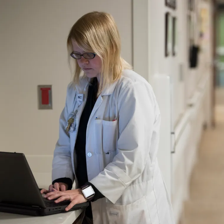 A Novant Health doctor is standing in a hallway typing on a laptop. 