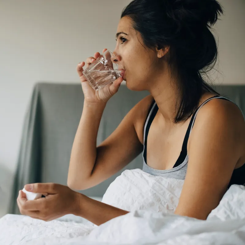 Woman sitting in bed drinking a glass of water as she takes medication. 