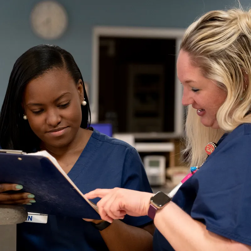 One Novant Health nurse is holding and reading information on a clip board. Another nurse is standing next to her as she looks and points at the information. 