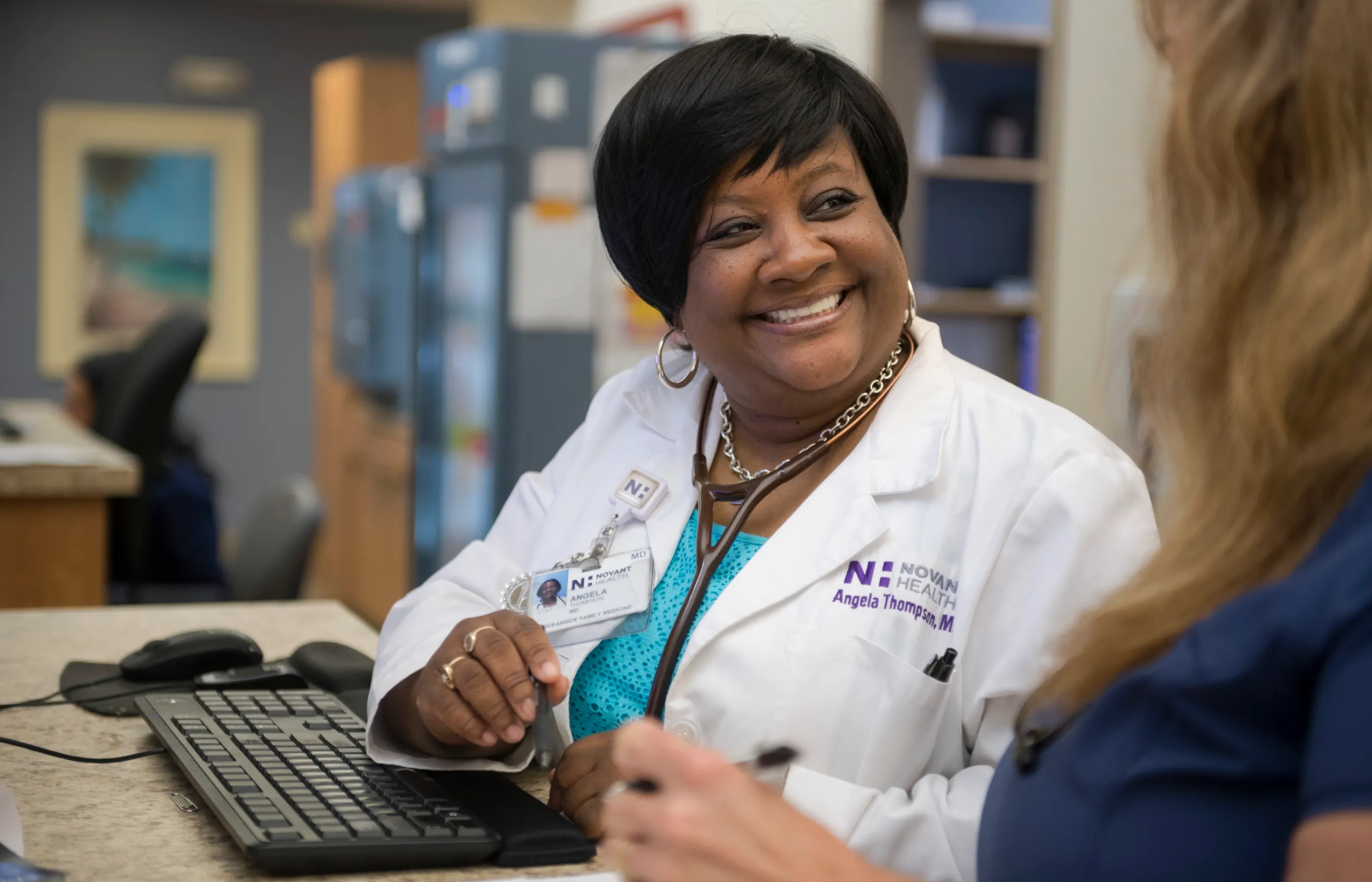 Novant Health Physician, Dr. Thompson, is sitting at a computer talking and smiling with a team member. 