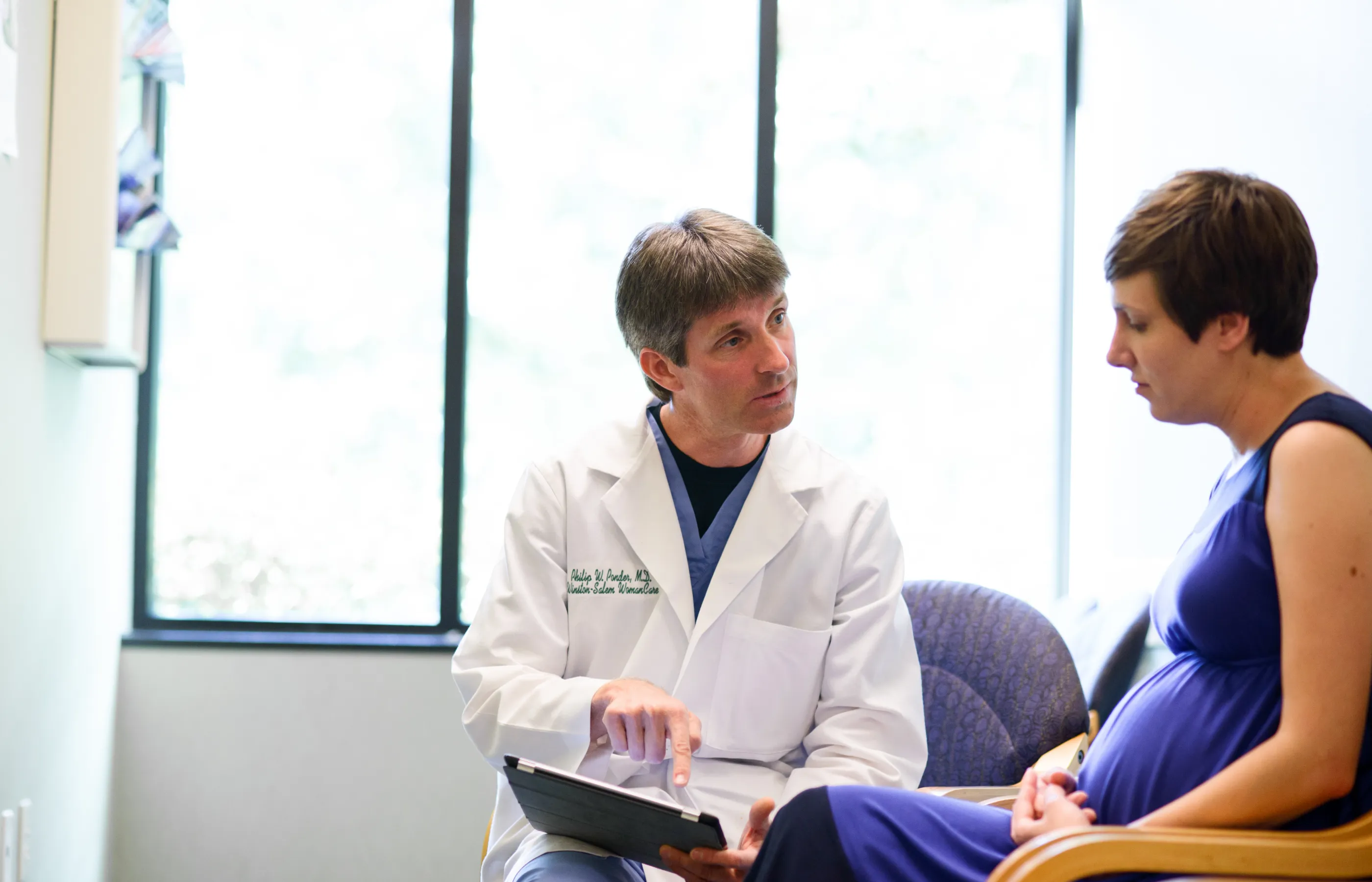 Dr. Ponder is sitting with a pregnant patient and reviewing information on a smart tablet. He is pointing at the tablet as he talks. The patient is looking and following along. 