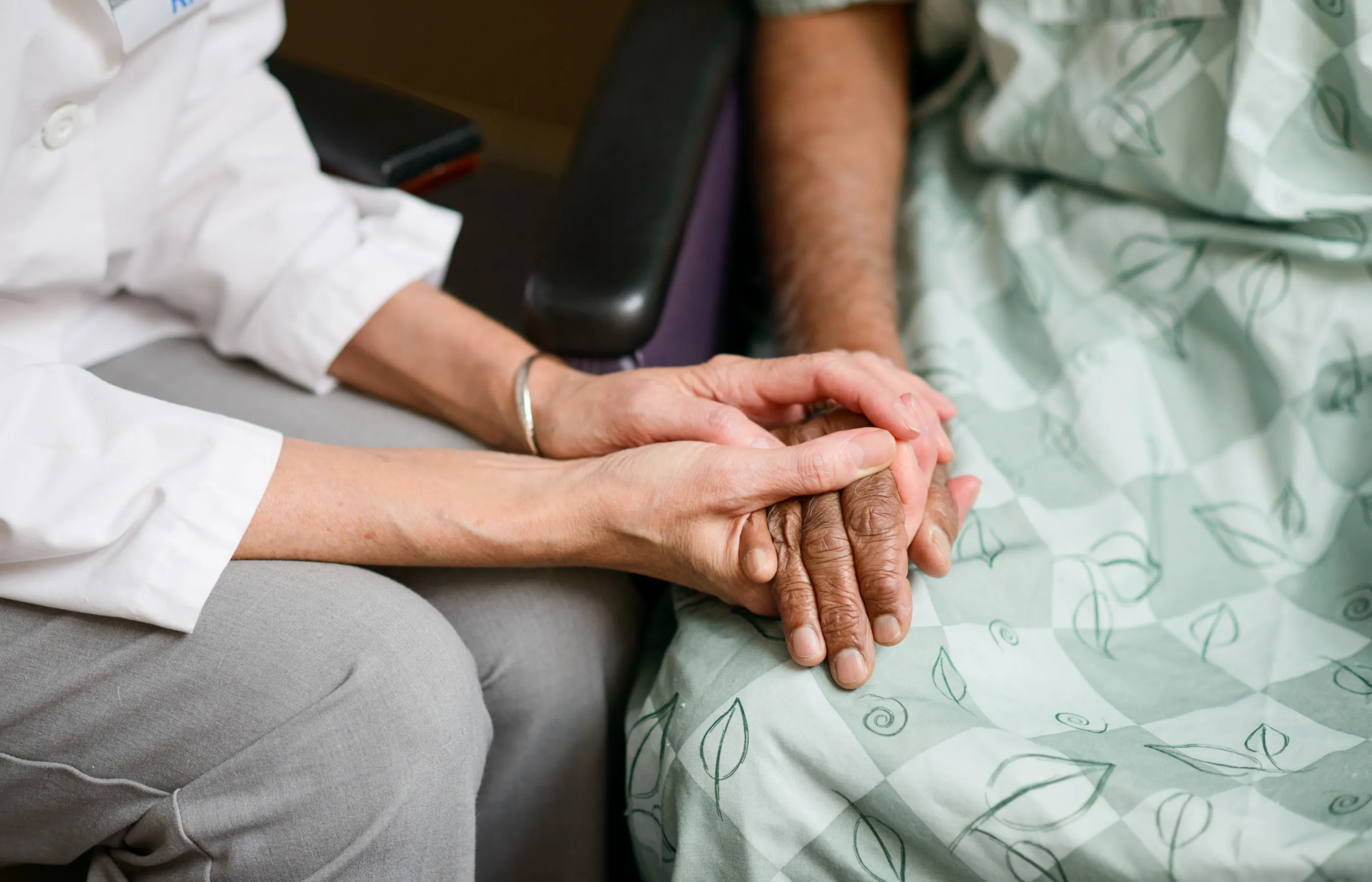 A doctor is sitting next to a patient, who has on a hospital gown. The doctor has grabbed a hold of resting the patients in support. 