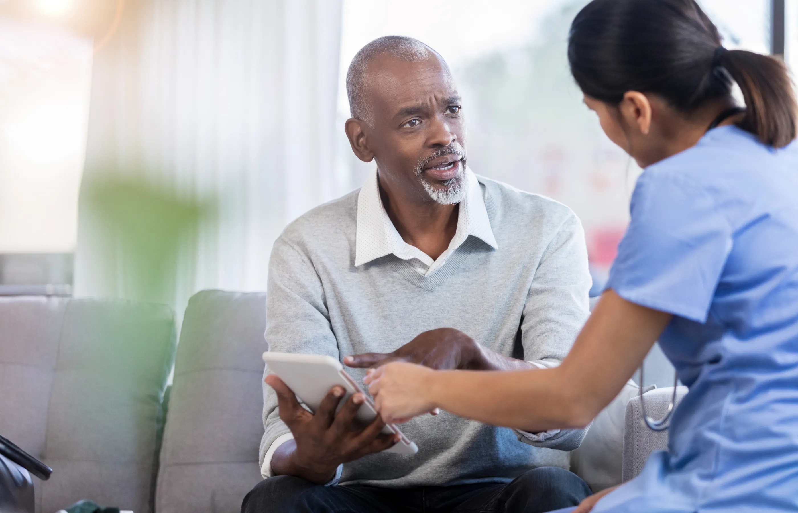 A male patient is sitting and talking with a nurse. Together, they are reviewing information on a tablet.