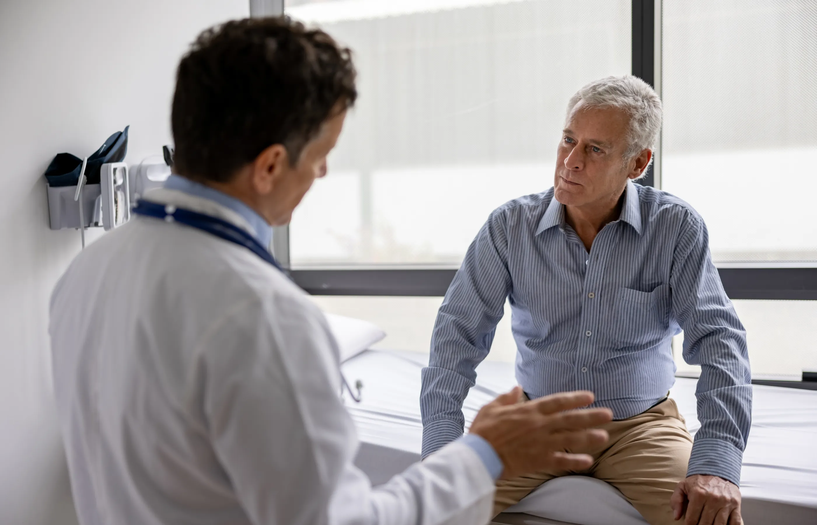 A mature man is sitting on an exam table talking with a doctor. 