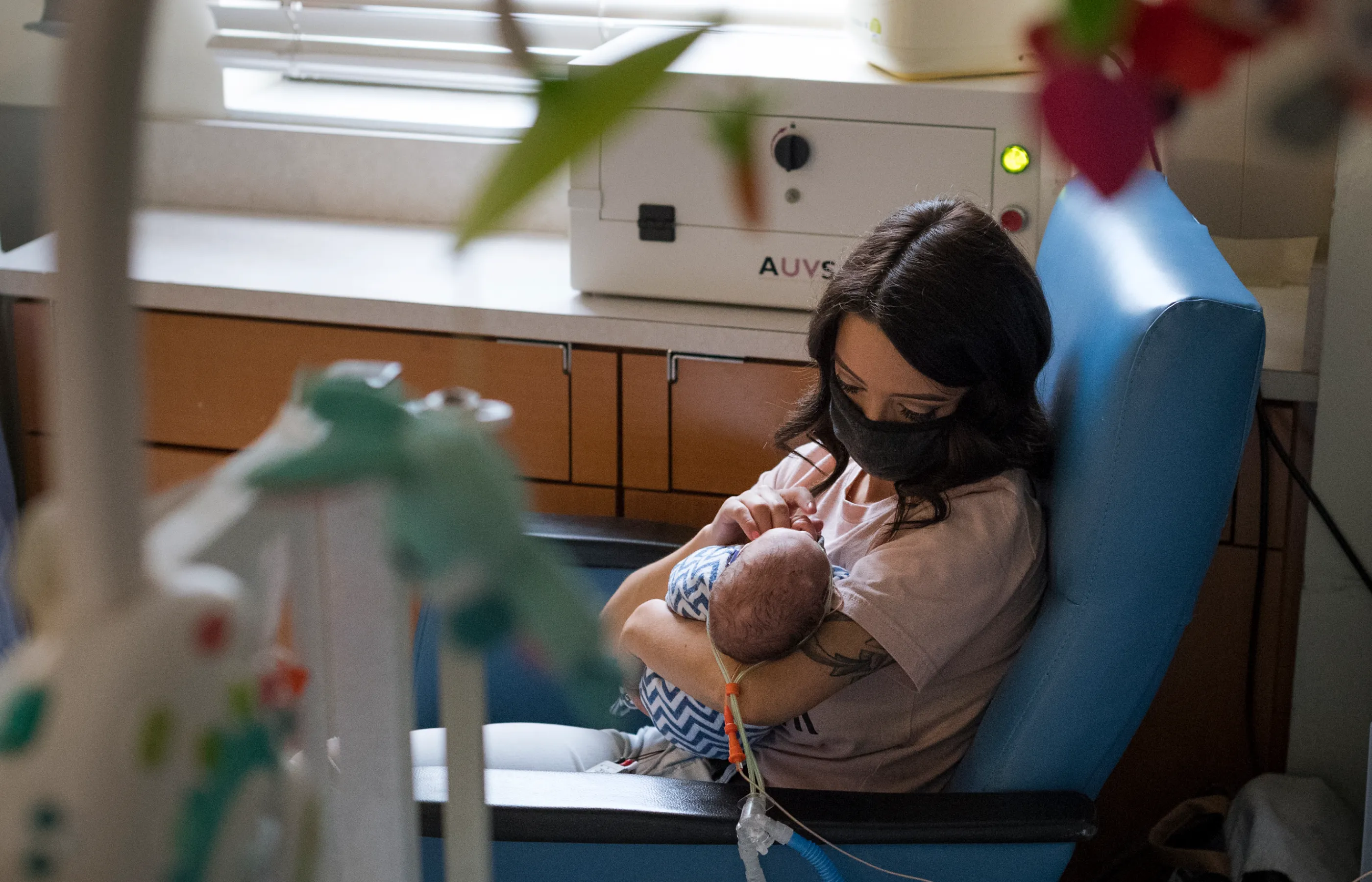 Mother is wearing a mask as she sits in a chair inside her babies NICU room. The child's bed mobile is in view as she is holds and rocks the baby. 