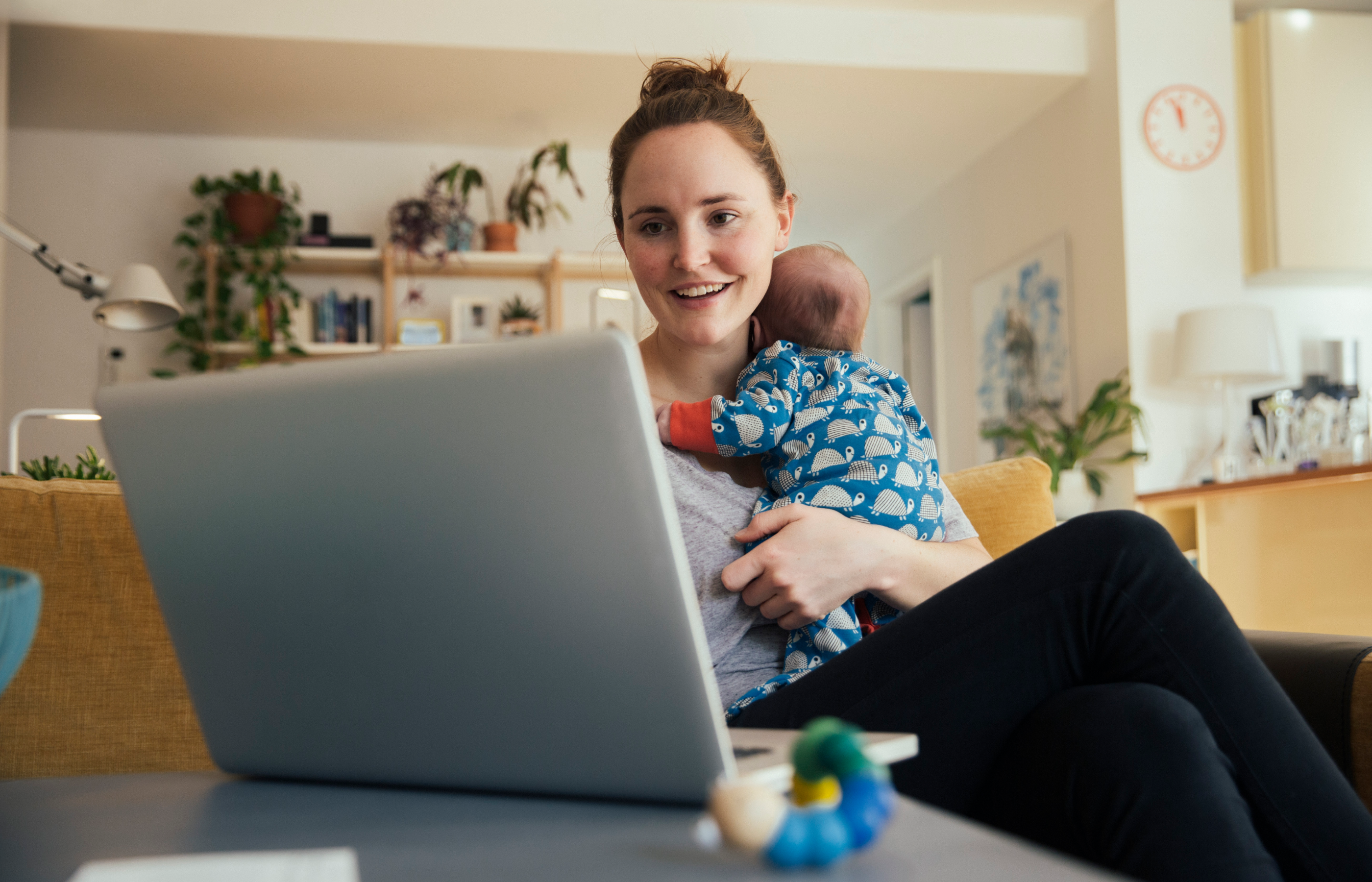 A mother is sitting on her couch holding her baby while looking at a laptop. A mother is sitting on her couch holding her baby while looking at a laptop.