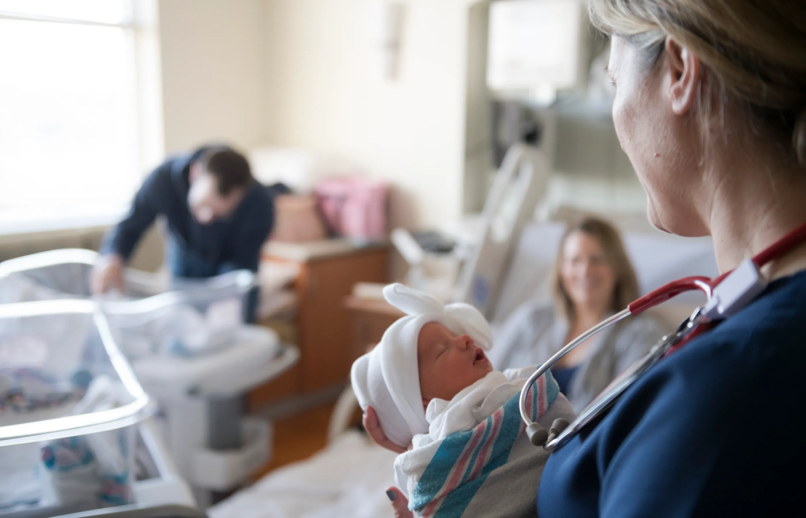 A nurse is in a hospital room, holding a newborn, as she checks in with newly born twins and their family. 