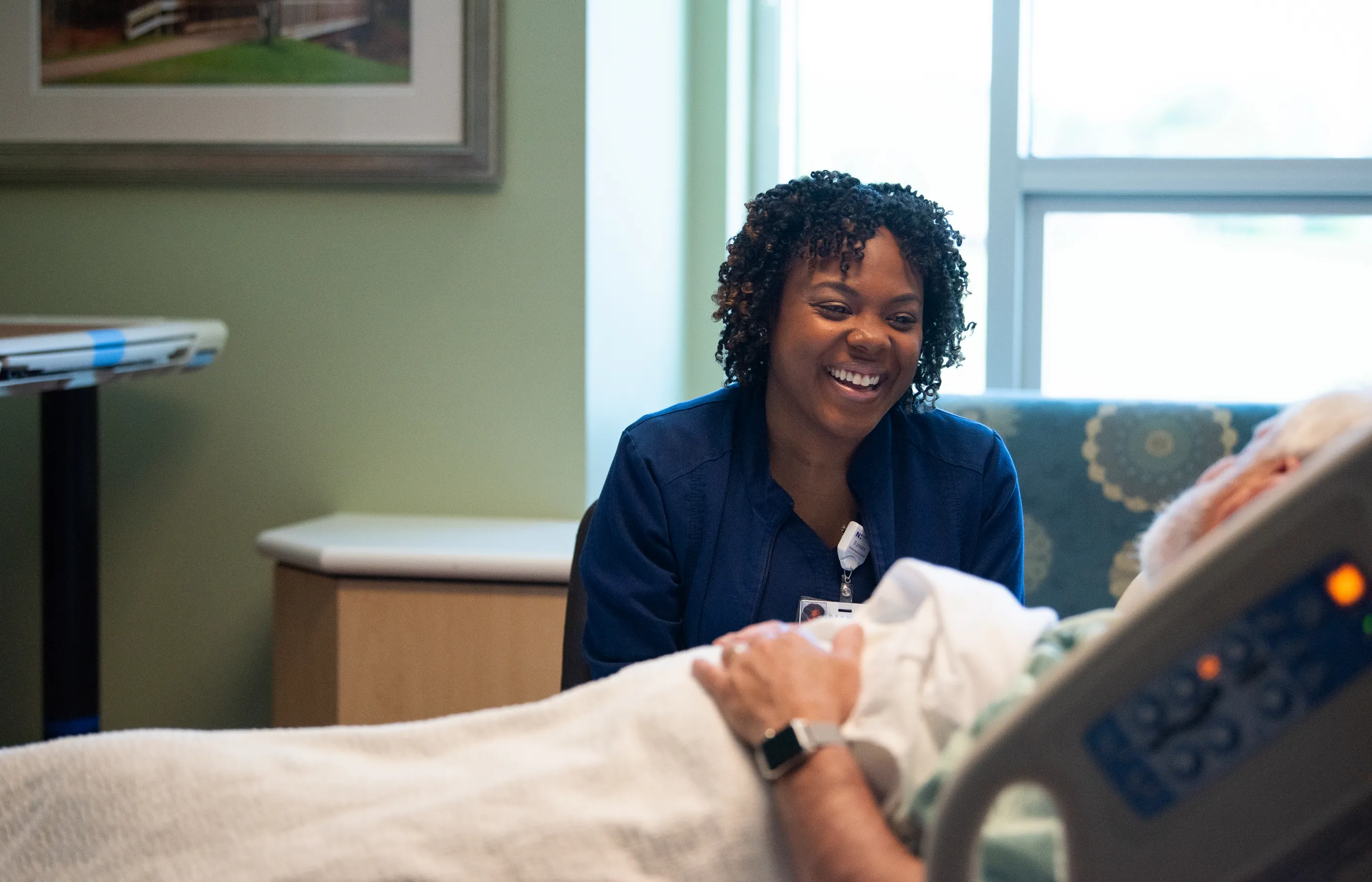 Novant Health nurse is sitting in a patients recovery room. The nurse is smiling and talking with the patient as they lie in bed.