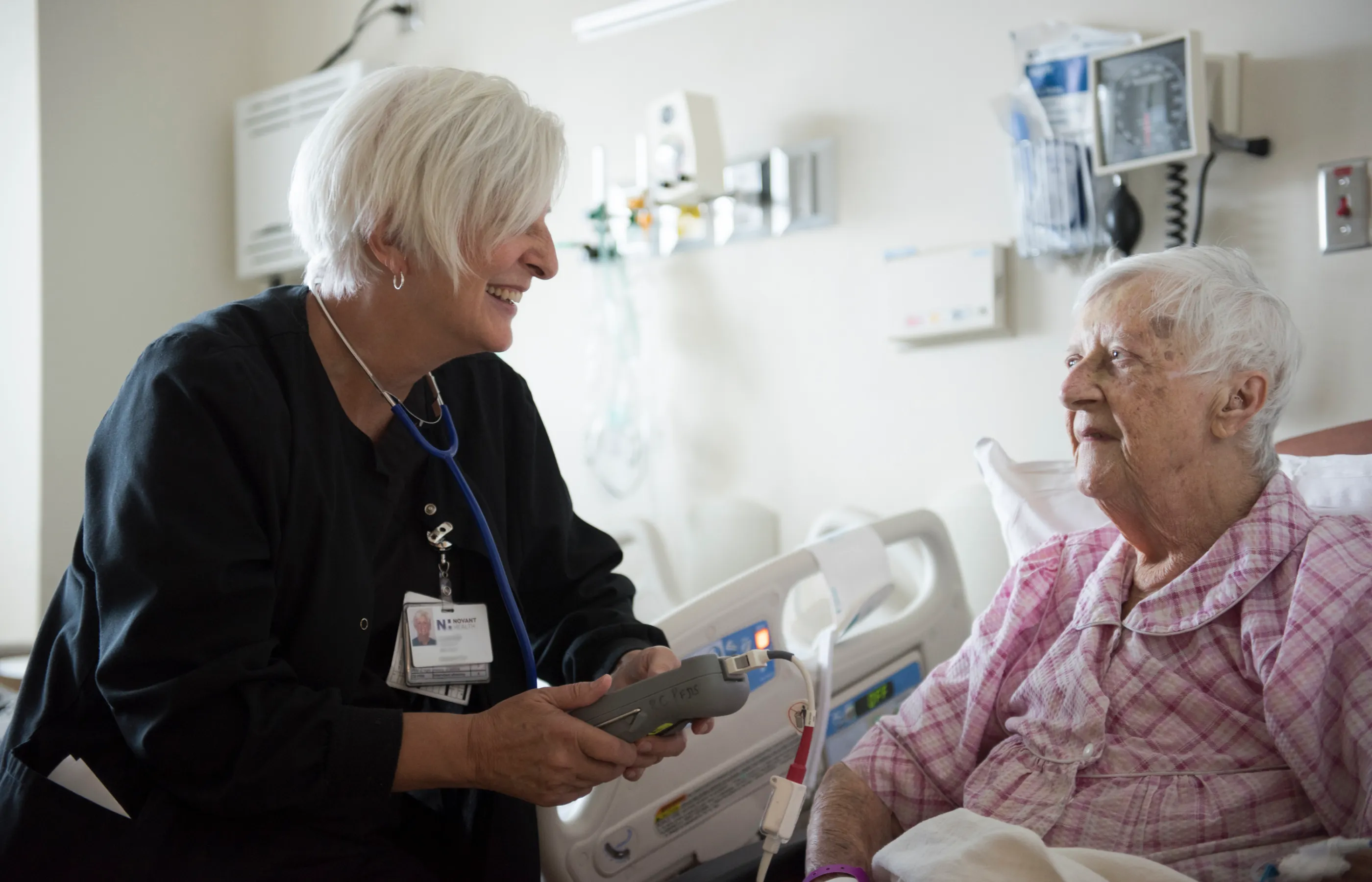A Novant Health nurse is at the bedside of a senior patient. The nurse is smiling and talking with the patient as she holders a large medical devce in her hand.