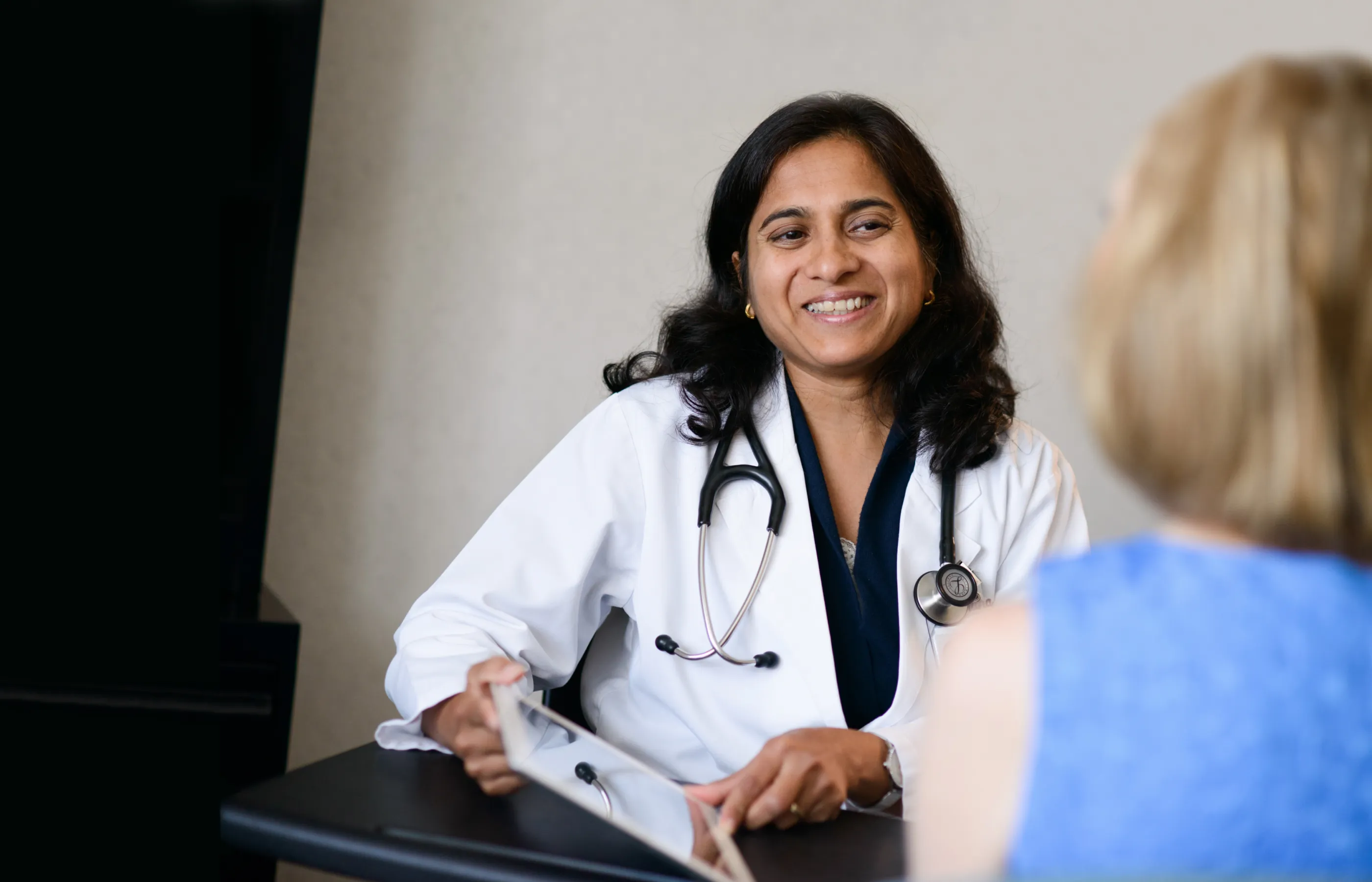 A physician is sitting with a lab coat and stethoscope around her neck as she talks with a patient. 