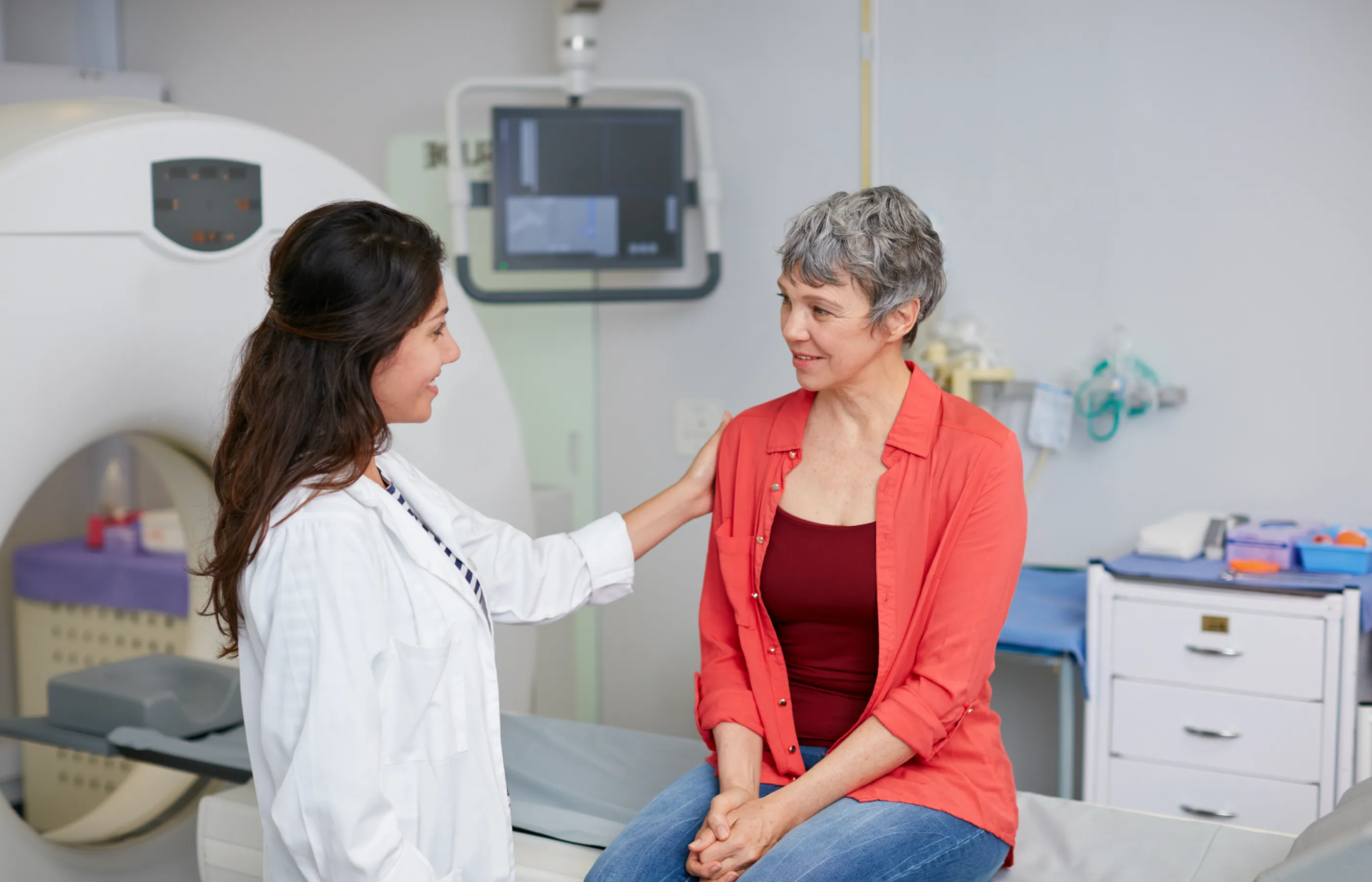 A healthcare provider is resting her hand on a patients shoulder as she is talking and preparing her for an imaging scan. A healthcare provider is resting her hand on a patients shoulder as she is talking and preparing her for an imaging scan.